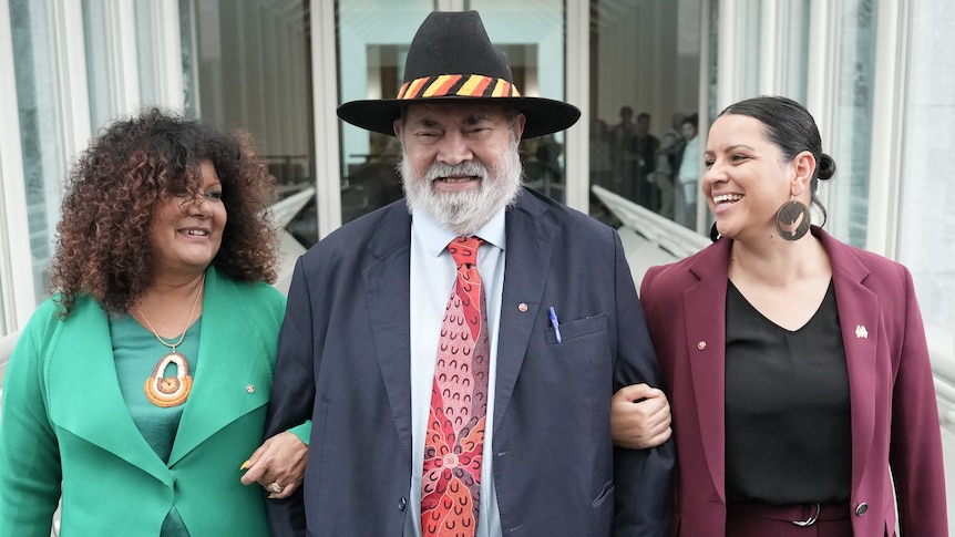 An older Aboriginal man wearing a suit and wide brimmed hat walks with two Indigenous women on either side of him.