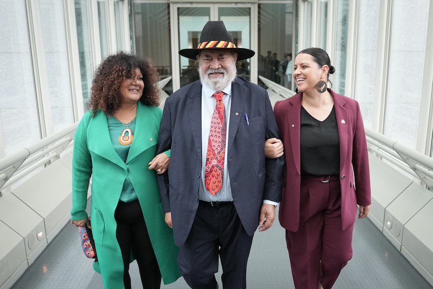 An older Aboriginal man wearing a suit and wide brimmed hat walks with two Indigenous women on either side of him.