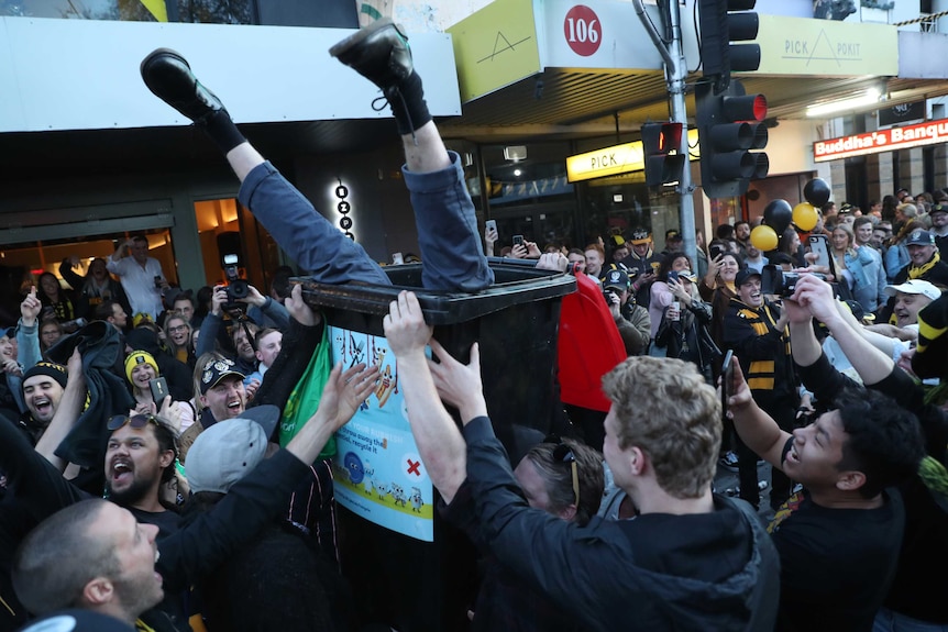 a person's legs stick out from a rubbish bin as tigers fans celebrate near a traffic light.