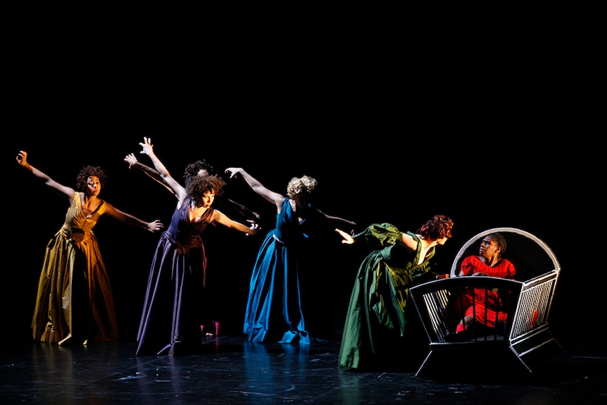 Five female dancers in dresses stand in line formation facing a woman in red dress seated in baby's crib on dark stage.