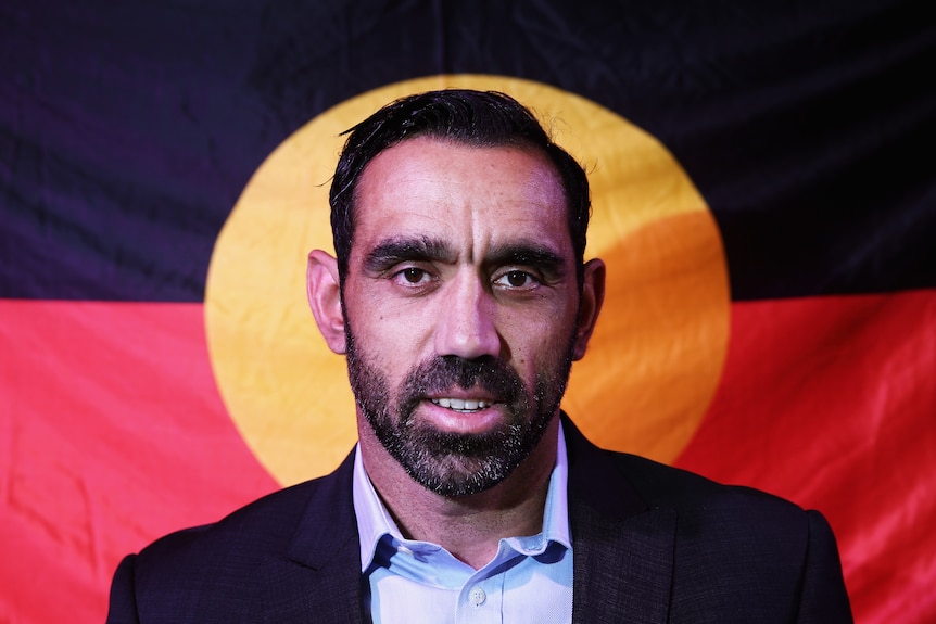Adam Goodes in a suit in front of the Aboriginal flag.