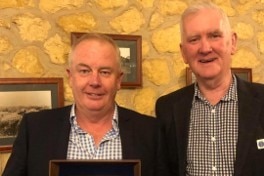 Two men smile at the camera, one holding a framed award