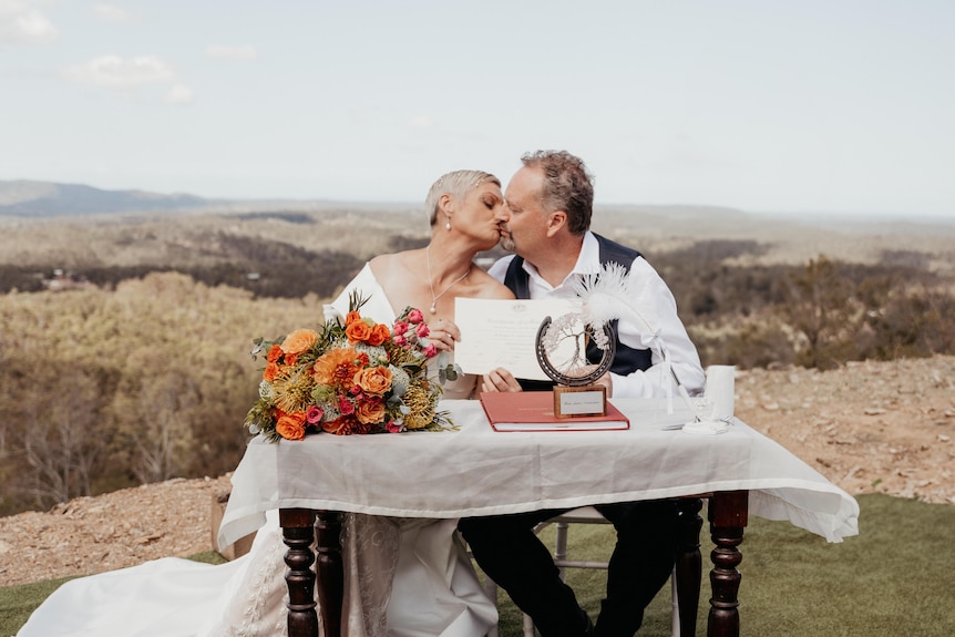 A married couple kiss at a table at their outdoor wedding.