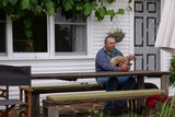 Man sits at a table on his verandah with a dog in his lap, looking at his phone.
