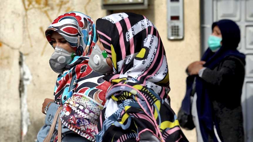 Women walk down the street wearing head scarves and masks.