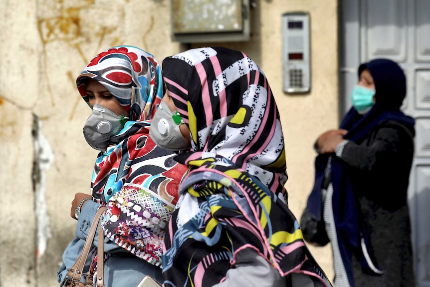 Women walk down the street wearing head scarves and masks.