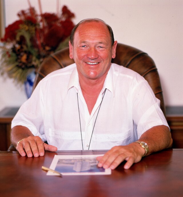 A smiling man in White shirt sits at a desk.