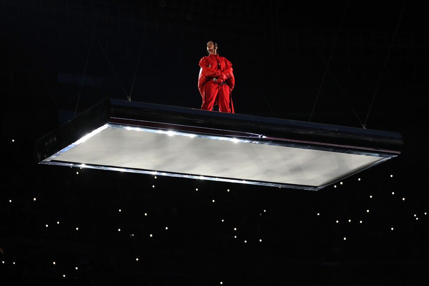Rihanna looks on as she stands on a wire-suspended white platform surrounded by darkness