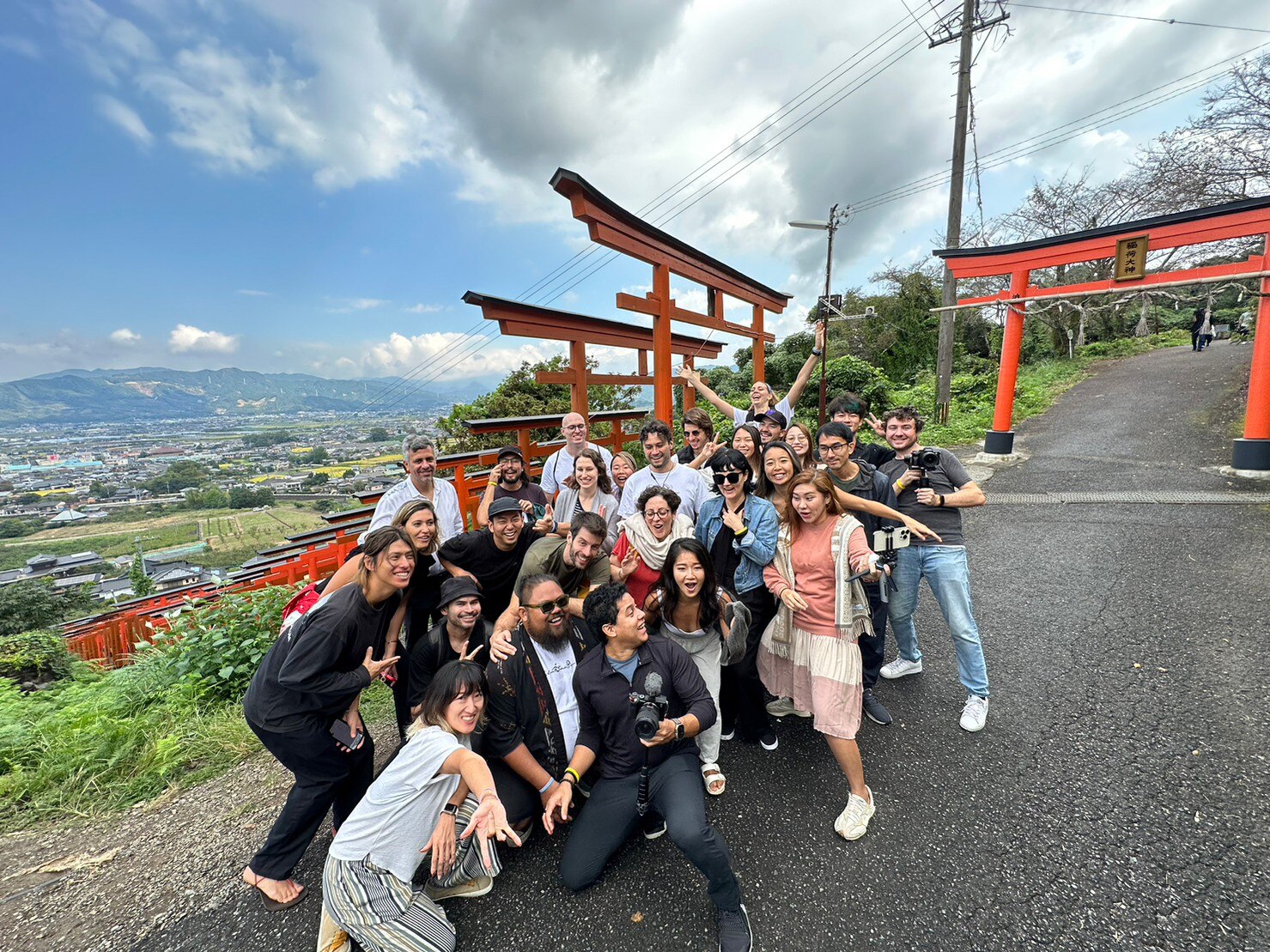 A group of smiling young folk pose for a fun group shot near orange and black torii gates on a hill in rural Japan.