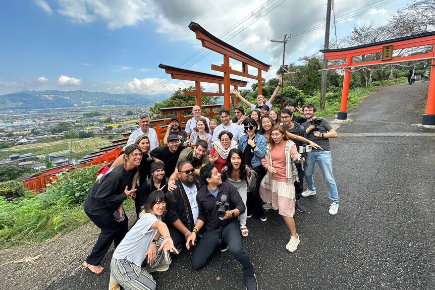 A group of smiling young folk pose for a fun group shot near orange and black torii gates on a hill in rural Japan.
