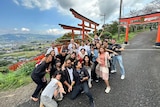 A group of smiling young folk pose for a fun group shot near orange and black torii gates on a hill in rural Japan.