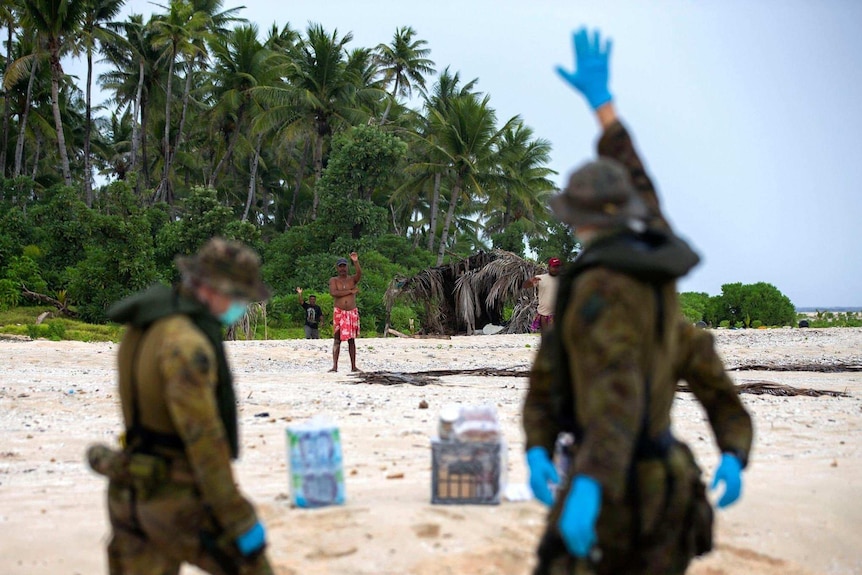 Two military personnel, in the foreground, wave to a bare-chested villager on a tropical island.