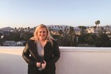 Rebel Wilson wearing a black leather jacket standing on a rooftop balcony with Sunset Boulevard behind her