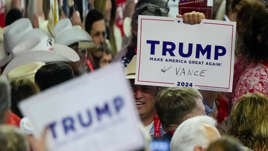 A person holds up a TRUMP campaign sign from a crowd, it has + VANCE written on in black pen
