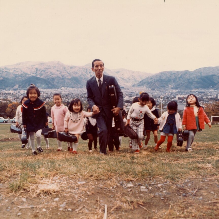 A colour photo shows Suzuki in a suit leading children carrying violins through the mountains
