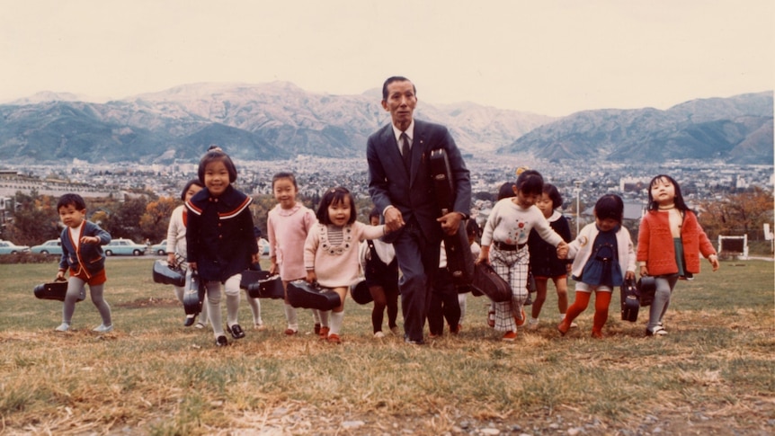 A colour photo shows Suzuki in a suit leading children carrying violins through the mountains