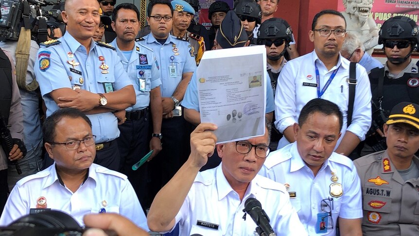 A man wearing glasses holds up stapled paperwork in his right hand while surrounded by uniformed officials and armed officers.