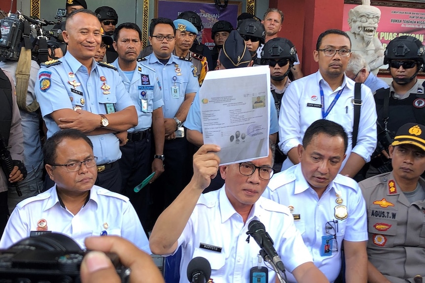 A man wearing glasses holds up stapled paperwork in his right hand while surrounded by uniformed officials and armed officers.