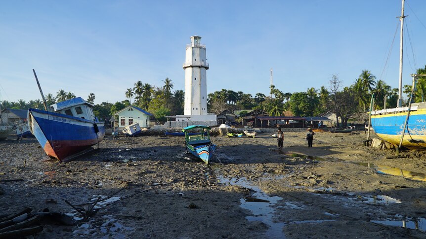 A small, blue fishing boat on the brown sandy shore, where a white lighthouse stands tall in the background. 