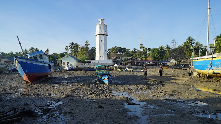 A small, blue fishing boat on the brown sandy shore, where a white lighthouse stands tall in the background. 