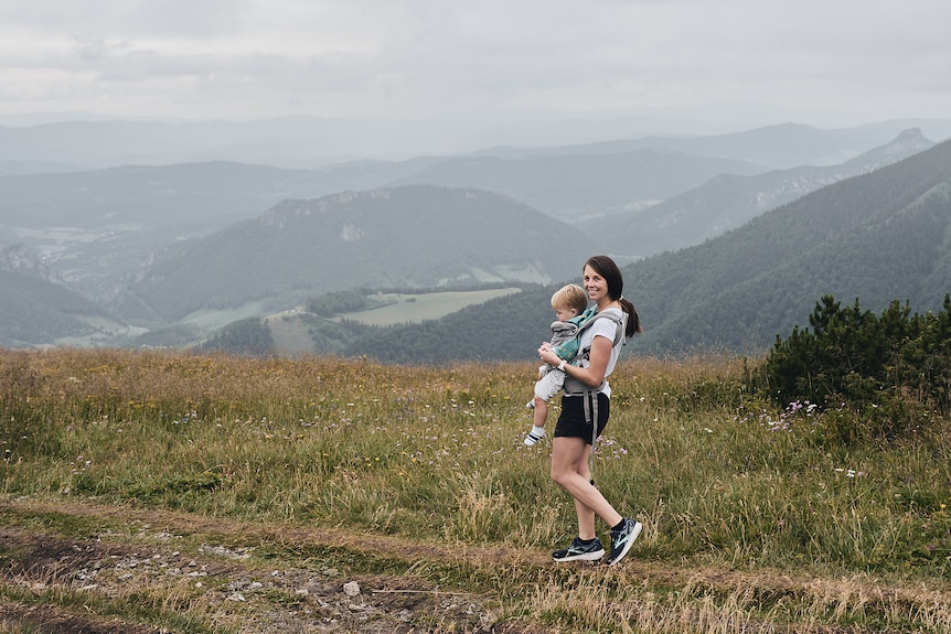Danielle smiles as she holds her son in a baby carrier while walking near some mountains.