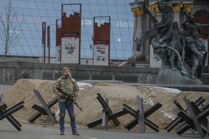 A man stands in the middle of the road, near a patriotic statue. There are barricades behind him.