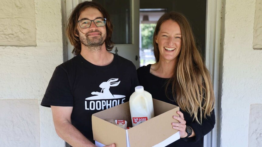 A man and a woman hold a box full of dairy products in front of an open door