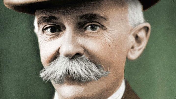 A colourised picture of a bushy- moustached man wearing a jacket, tie and homburg hat.
