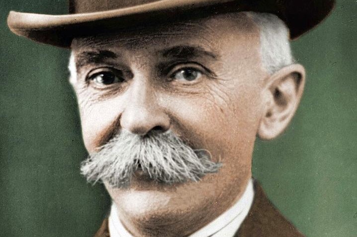 A colourised picture of a bushy- moustached man wearing a jacket, tie and homburg hat.