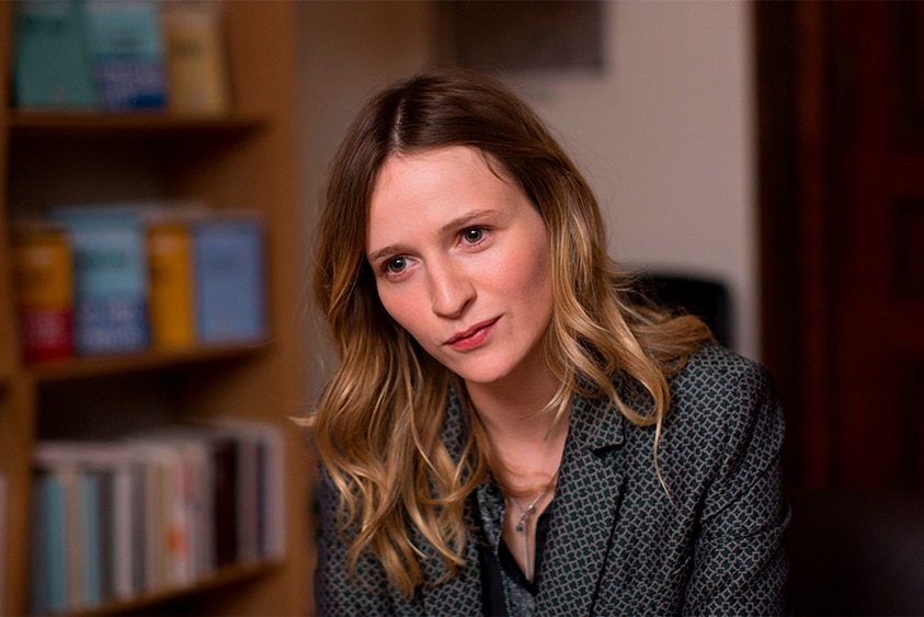 A young woman with strawberry blond hair wears dark patterned blazer and sits in interior room near bookcase.