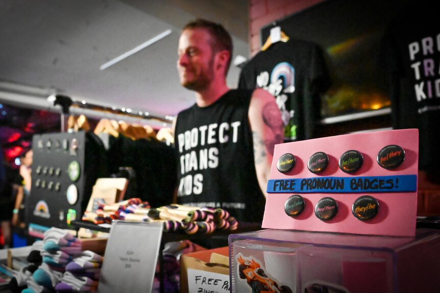 A person stands behind a table of merch, their shirt reads 'protect trans kids''