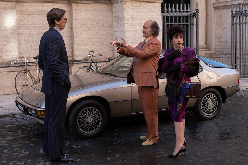 Man in navy suit with hands in pockets stands in front of balding man in checked orange suit and woman in vivid purple dress.