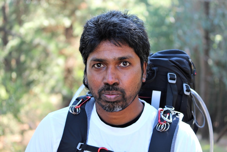 A close-up of a Sri Lankan man's face, wearing a backpack, looking directly at the camera