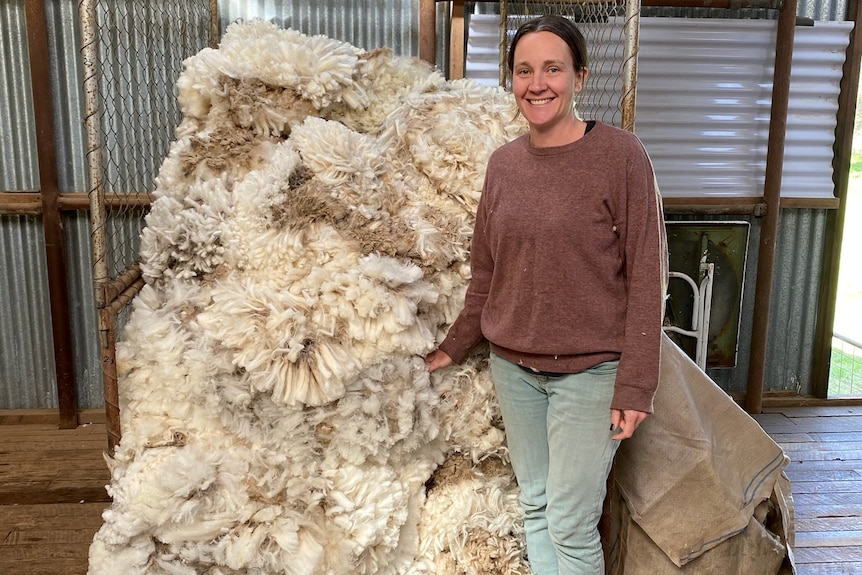 A fair-skinned female farmer, Verity, stands smiling next to a large bale of wool in a tin shearing shed. 