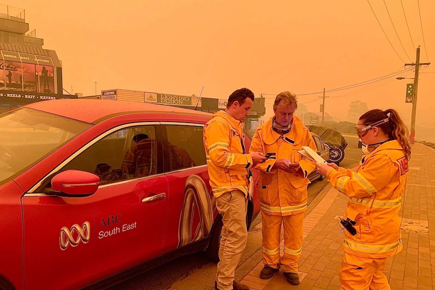 Three people standing around car looking at phones and notebook with smoky orange sky.