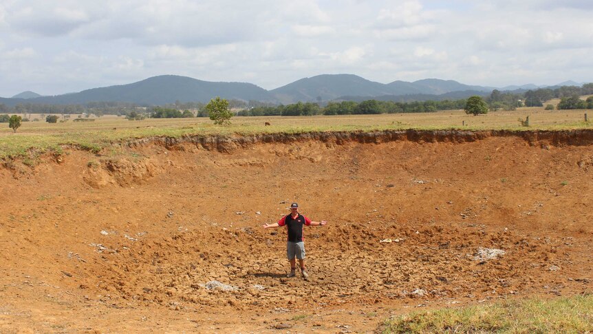 A man stands in the middle of a dry dam, surrounded by brown farmland.