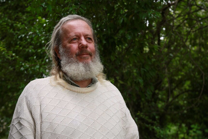 A man with a big white beard wears a knitted jumper in front of trees.