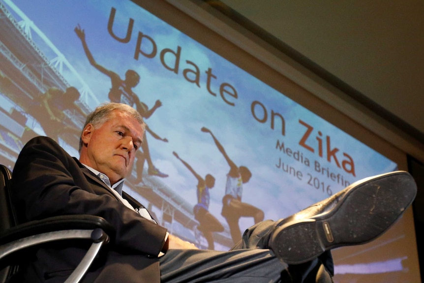 A man sits looking down, behind is a screen with pictures of athletes and the sign "Update on Zika".