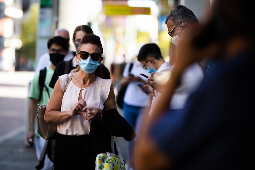 Woman with mask and sunglasses on walking past other people with masks on waiting for a bus.