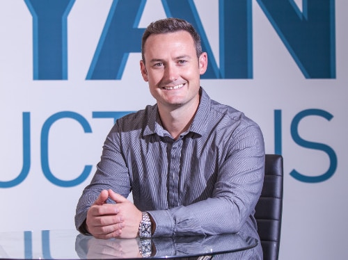 A middle aged white man sits at a glass table with his hands clasped together smiling at the camera.