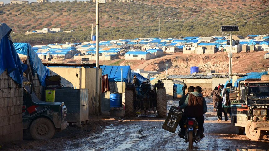 Two men ride a motorbike through a refugee camp of makeshift brick houses with blue tarpaulin roofs