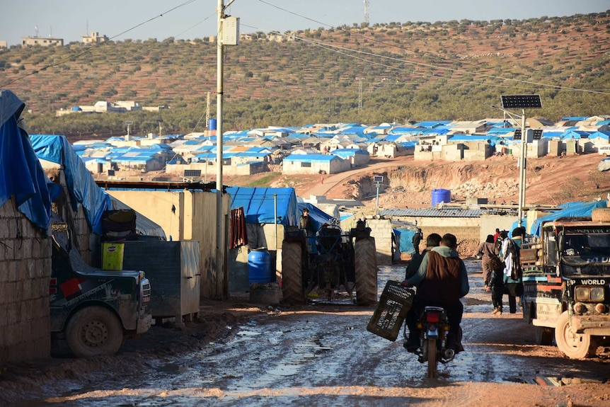Two men ride a motorbike through a refugee camp of makeshift brick houses with blue tarpaulin roofs