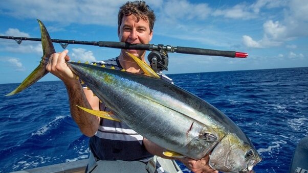 Clarke Gayford stands in a boat at sea with a fishing rod in his mouth and a live fish in his hands.