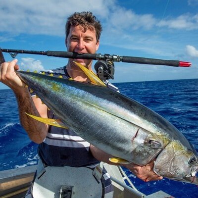Clarke Gayford stands in a boat at sea with a fishing rod in his mouth and a live fish in his hands.