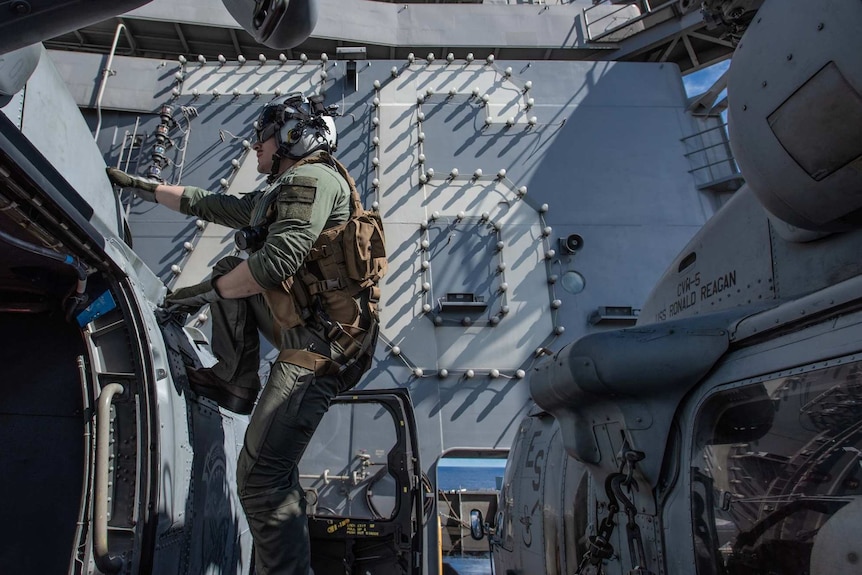 A US navy man climbs a ladder on the ship, behind him is an aircraft.