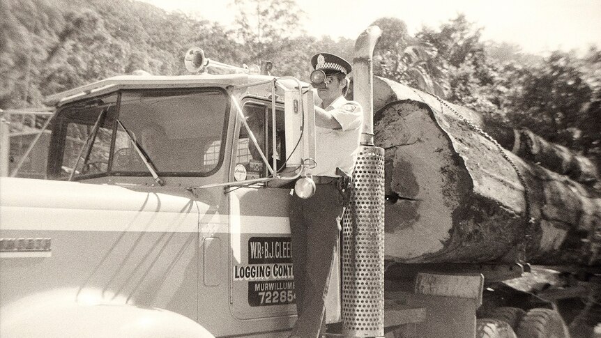 A black and white photo of a police officer standing on the side of a logging truck.
