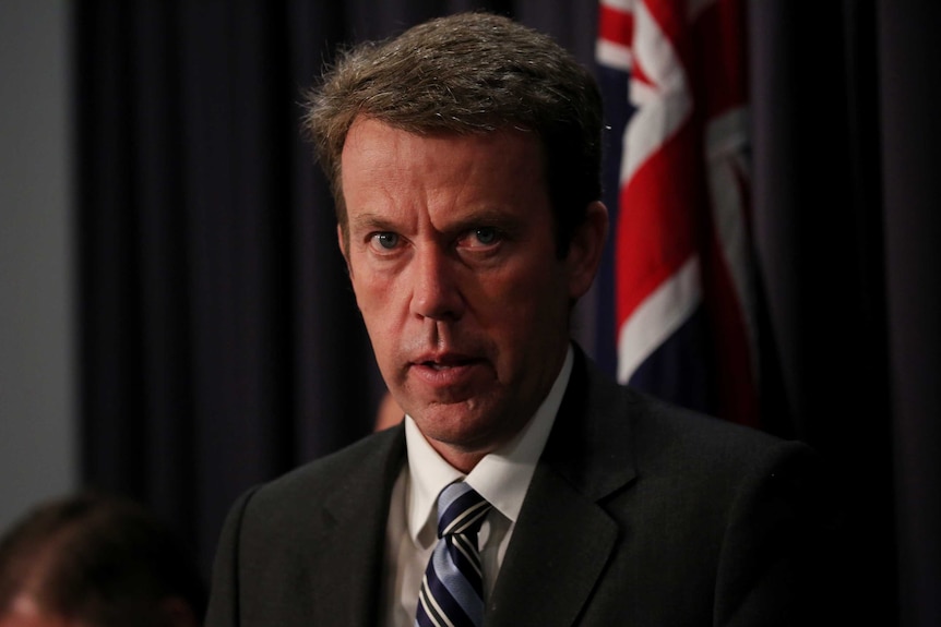 Dan Tehan looks at the camera. An Australian flag is in the background, and he's wearing a blue striped tie.