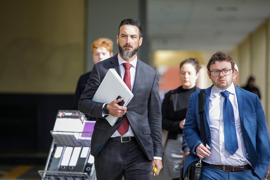 Two men in suits walk towards the camera, holding folders and bags.