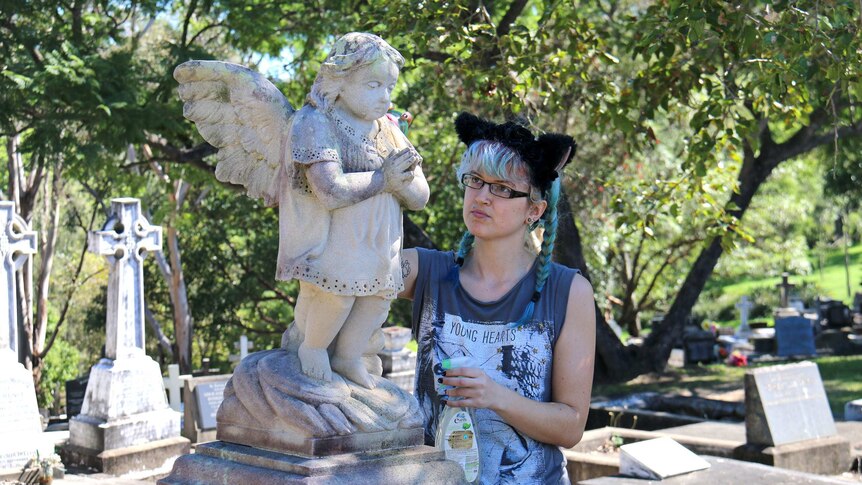 A volunteer cleans a statue of an angel on a headstone at South Brisbane cemetery.