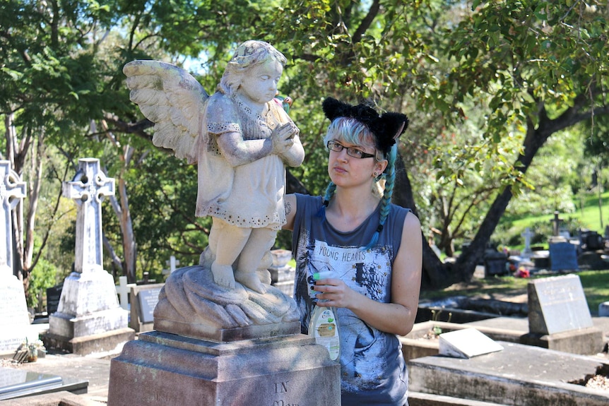 A volunteer cleans a statue of an angel on a headstone at South Brisbane cemetery.